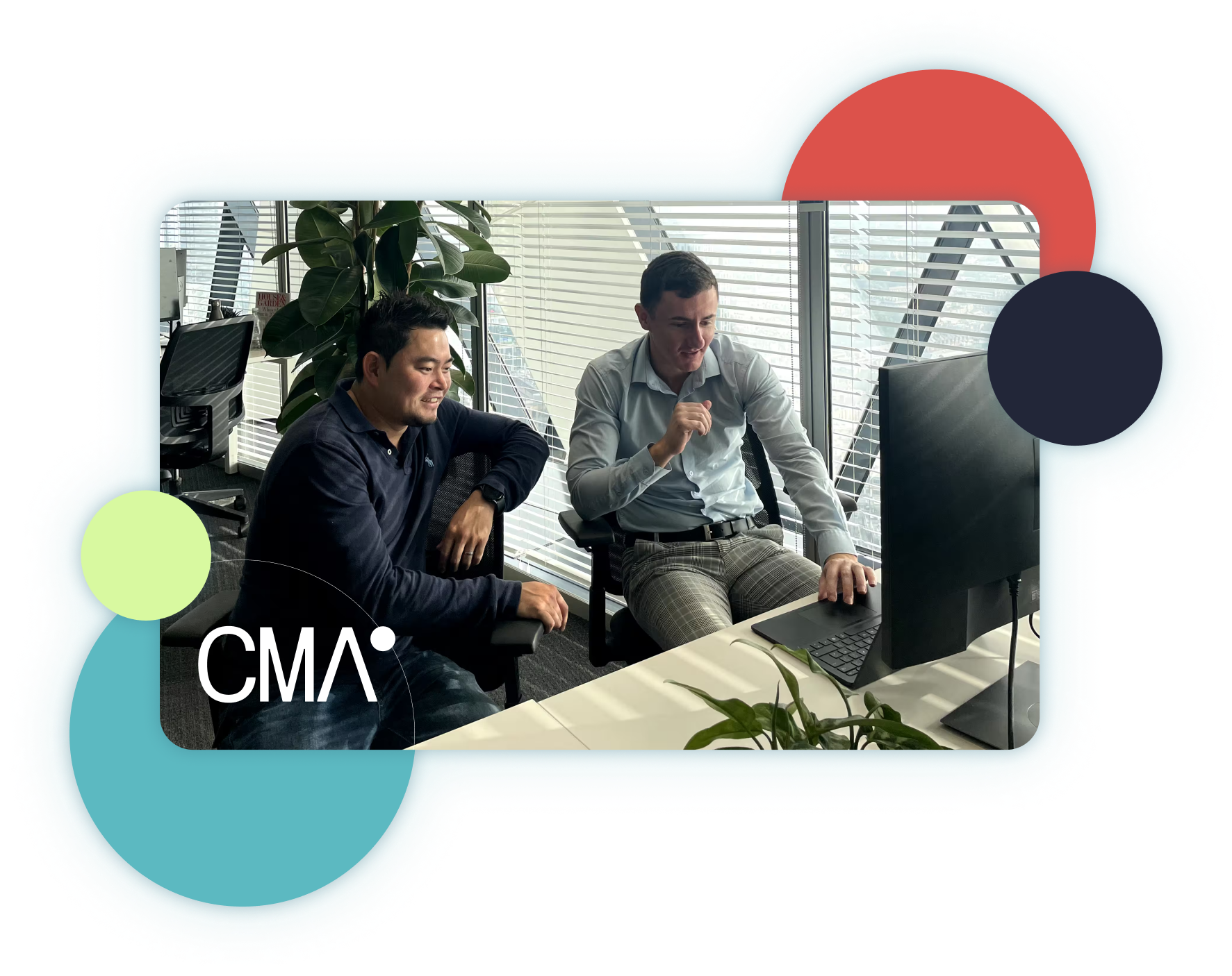 Two men sitting at a desk in front of a computer monitor, engaged in a discussion in an office with large windows and blinds.