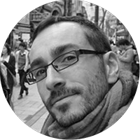 Close-up black and white photo of a man with glasses, short hair, and a scarf, with a city street and pedestrians in the background.